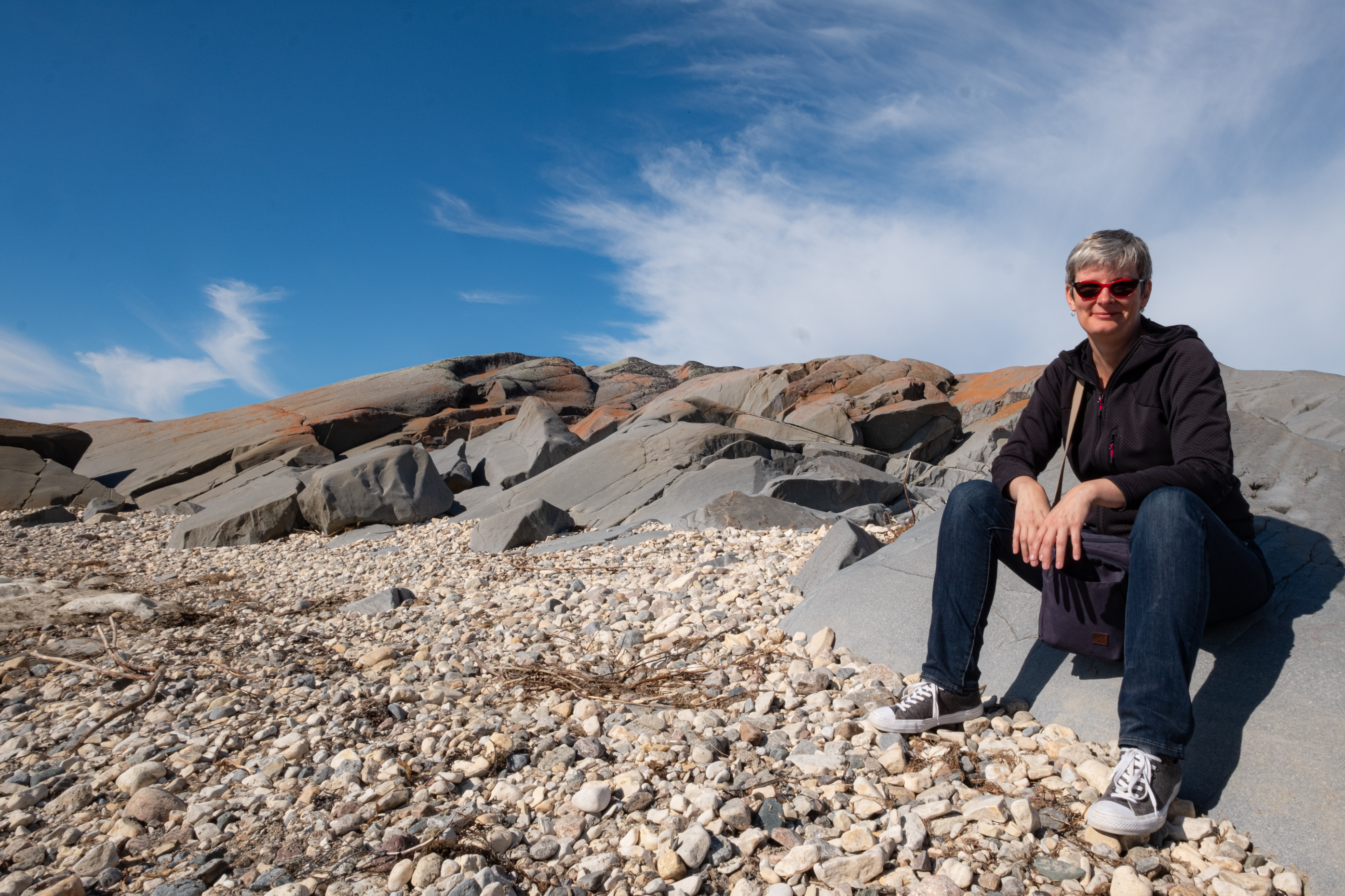 A woman with short grey hair wearing glasses smiles while sitting on a rocky landscape in northern Manitoba.
