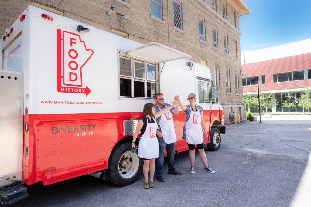 Three people (Kimberley Moore, Kent Davies, and Janis Thiessen) wearing white aprons with the Manitoba Food History Project logo give each other a high five while standing in front of a red and white food truck on a warm summer day on the University of Winnipeg campus.
